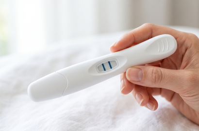 White home pregnancy test showing two blue lines, held in hand on a bed.