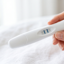 Close-up of a hand holding a digital thermometer on a white bed sheet, showing a temperature reading on the display.